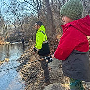 Colson is registered to the contest to win money with this photo: child, adult, fishing, creek, riverbank, rock, winter_clothing, knit_hat, jacket, boots, trees, leafless_trees, outdoor, nature, water, fishing_rod, person, daylight, recreation, forest