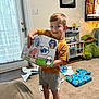 Colson is registered to the contest to win money with this photo: child, boy, smiling, toy_box, living_room, carpet, barefoot, shorts, tshirt, present, wrapping_paper, gift, plush_toy, stuffed_animal, play_kitchen, television, mirror, door, light_switch, home_interior