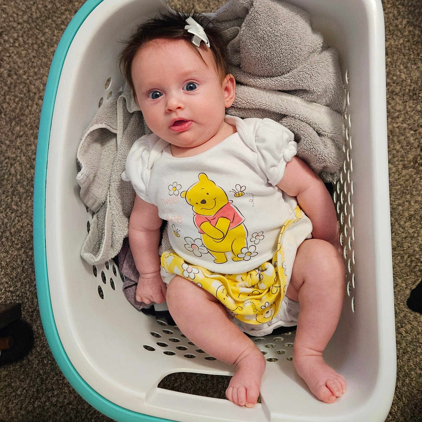 Winslow is registered to the contest to win money with this photo: baby, laundry_basket, towel, carpet, clothing, winnie_the_pooh, infant, hair_bow, cute, indoors, child, person, relaxing, soft_texture, white_basket, yellow_clothing, portrait, smiling, happy, comfortable