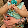 baby, pacifier, tattoo, adult_hand, outdoor, tree, greenery, child, infant, holding, summer_clothing, cute, portrait, skin, person, nature, casual, daylight, expression, sitting