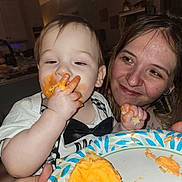 Oliver joined the competition — help win amazing prizes! baby, bow_tie, celebration, child, clothing, cupcake, eating, face, family, food, hand, happy, indoor, kitchen, messy, orange_frosting, person, plate, smile, woman