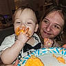 baby, bow_tie, celebration, child, clothing, cupcake, eating, face, family, food, hand, happy, indoor, kitchen, messy, orange_frosting, person, plate, smile, woman