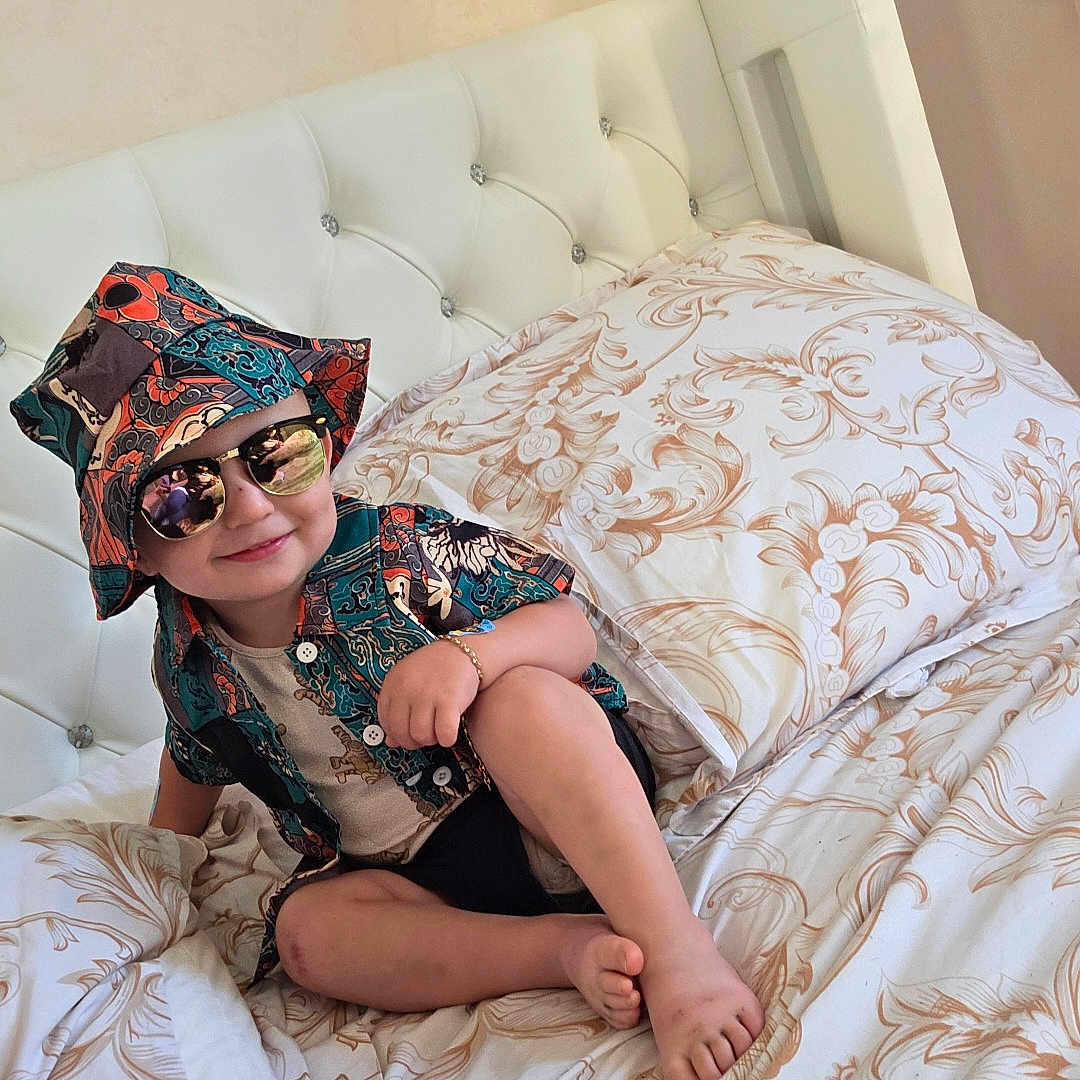 Eden a rejoint le concours — aidez-le/la à gagner de superbes lots ! barefoot, bed, bedding, bracelet, bucket_hat, child, cute, fashion, hat, headwear, indoor, patterned_clothing, person, pillow, portrait, relaxed, shirt, smile, sunglasses, toddler