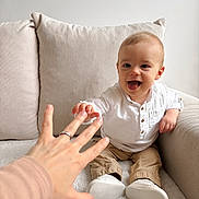 Noé a rejoint le concours — aidez-le/la à gagner de superbes lots ! adult_hand, baby, beige_pants, blanket, child, couch, cozy, curious, furniture, hand, happy, indoor, interaction, pillow, portrait, reaching, ring, smiling, white_shirt, white_shoes