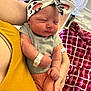 newborn, baby, infant, headband, cartoon_print, hospital_wristband, adult_arm, holding, sleeping, cozy_clothing, red_blanket, fan, indoor, close_up, person, skin, hand, face, peaceful, nursery
