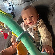 Sandro participe au concours pour gagner de l'argent avec cette photo : baby, bib, blanket, bouncer, carpet, clothing, coffee_table, couch, curtain, eyes, hand, infant, living_room, play_toy, remote_control, smiling, straps, sunlight, toy_bar, wooden_button