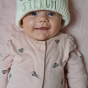 Jessie a rejoint le concours — aidez-le/la à gagner de superbes lots ! baby, cartoon_prints, child, clothing, cute, face, happy, hat, indoors, infant, knit_hat, lying_down, person, pink_onesie, plush_surface, pom_pom, portrait, smiling, soft_texture, stitch_character