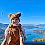 baby, child, person, hat, clothing, outdoor, sky, mountain, lake, water, nature, paraglider, hands, smile, scenery, blue_sky, forest, fall, sunny, portrait