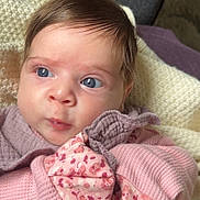 Leah is registered to the contest to win money with this photo: baby, infant, face, blue_eyes, pink_clothing, purple_clothing, blanket, knitted_blanket, soft_texture, closeup, portrait, cute, child, young, indoor, skin, hair, expression, curious, resting