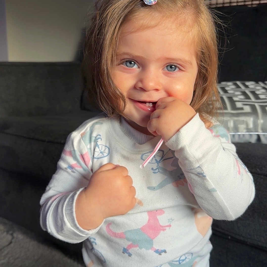 Leah joined the competition — help win amazing prizes! child, girl, toddler, portrait, smiling, happy, standing, pajamas, blonde_hair, hair_clip, blue_eyes, indoors, living_room, sofa, cushion, sunlight, natural_light, casual, playful, animal_print