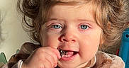 Leah joined the competition — help win amazing prizes! toddler, child, portrait, smiling, eating, snack, pigtails, curly_hair, blue_eyes, face, hand, clothing, jacket, indoors, toy, colorful, close_up, sunlight, happy, playful