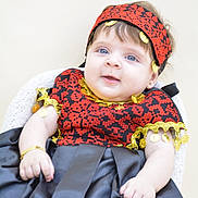 Leah is registered to the contest to win money with this photo: baby, child, infant, portrait, sitting, smiling, happy, face, eyes, headband, traditional_clothing, red, black, gold, jewelry, bracelet, earring, chair, indoor, close_up