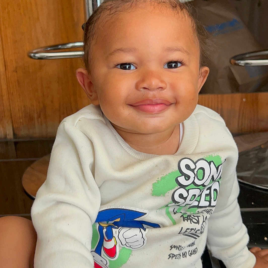 Ryan is registered to the contest to win money with this photo: toddler, child, smile, braided_hair, white_sweatshirt, sonic_the_hedgehog, graphic_tee, sitting, floor, tile_floor, wooden_background, indoor, happy, cute, portrait, young_child, casual, clothing, person, face