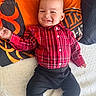Kingston is registered to the contest to win money with this photo: baby, child, smile, plaid_shirt, red_shirt, black_pants, socks, pillow, orange, black, white_background, indoor, happy, cute, face, person, portrait, fabric, texture, laying