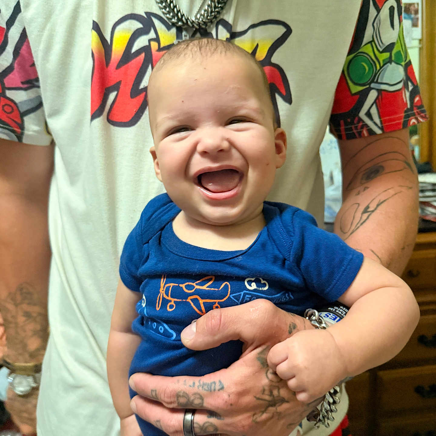 Kingston is registered to the contest to win money with this photo: baby, child, smiling, laughing, blue_onesie, tattooed_hand, adult, indoor, person, holding, happy, joyful, closeup, portrait, colorful_clothing, furniture, wooden_drawer, ring, chain, casual_wear