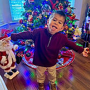 Jaxson is registered to the contest to win money with this photo: child, christmas_tree, holiday_decorations, tongue_out, playful, indoor, festive, lights, ornaments, santa_figurine, wood_floor, window, toy, smiling, person, celebration, red_clothing, beige_pants, brown_shoes, home