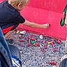 child, blond_hair, black_shirt, red_wall, gravel, colorful_objects, crouching, outdoor, person, footwear, casual, reaching, ground, side_view, young, curious, daylight, activity, clothing, texture
