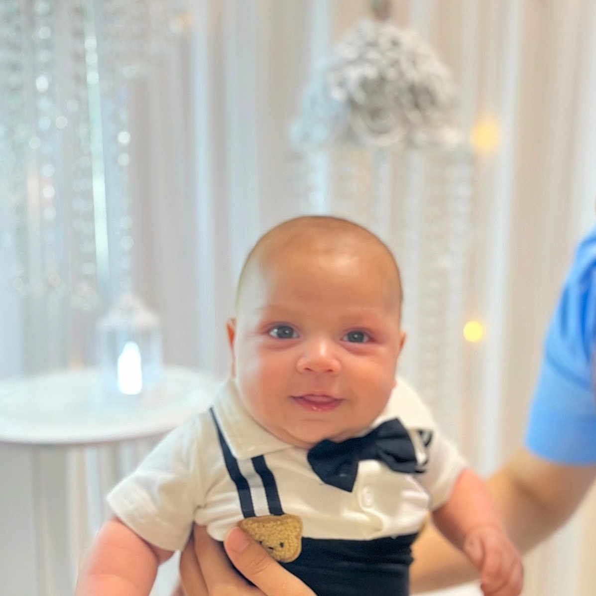 Eithan Moncada joined the competition — help win amazing prizes! adult_hands, baby, blue_eyes, bow_tie, child, close_up, curtains, decor, family_photo, formal_clothing, held, home, indoors, infant, portrait, smiling, suspenders, table, warm_lighting, window