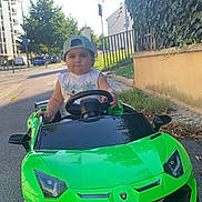 Yanis a rejoint le concours — aidez-le/la à gagner de superbes lots ! child, toddler, toy_car, green_car, outdoor, street, cap, steering_wheel, summer, sunlight, residential_area, sidewalk, trees, grass, building, play, fun, vehicle, cute, portrait