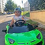 child, toddler, toy_car, green_car, outdoor, street, cap, steering_wheel, summer, sunlight, residential_area, sidewalk, trees, grass, building, play, fun, vehicle, cute, portrait