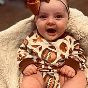 Gracie is registered to the contest to win money with this photo: baby, infant, smiling, happy, face, headband, bow, clothing, football_pattern, blanket, cozy, cute, child, skin, hand, legs, indoors, portrait, sitting, warm