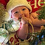 baby, santa_hat, christmas_light, holiday, festive, cheeks, cute, infant, decorations, christmas_tree, holiday_season, red, white, green, cozy, smile, cute_expression, indoors, soft_light, celebration