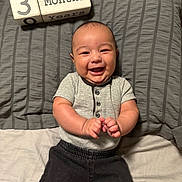 Jionni is registered to the contest to win money with this photo: baby, infant, smiling, clapping, milestone_blocks, 3_months, gray_pillow, bed, lying_down, short_sleeve_shirt, black_pants, happy, cute, portrait, child, young_baby, indoors, human, person, cheerful