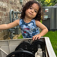 Jacob Sanders is registered to the contest to win money with this photo: child, curly_hair, tank_top, outdoor, brick_wall, utility_box, greenery, grass, summer, casual_pose, cute, smiling, person, daylight, playful, young_child, portrait, natural_light, happy, equipment