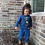 toddler, child, curly_hair, blue_sweatshirt, jeans, brown_boots, brick_wall, window, garden_bed, rocks, outdoor, walking, exploring, casual_clothing, person, daylight, curious, young_child, nature, sidewalk