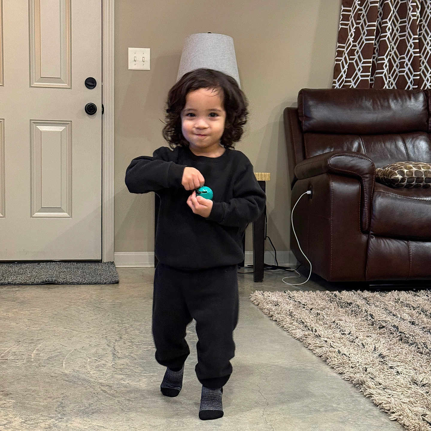 Jacob Sanders is registered to the contest to win money with this photo: black_clothing, carpet, child, concrete_floor, curly_hair, curtains, door, furniture, holding_object, home, indoor, lamp, leather_couch, living_room, person, rug, smiling, toddler, toy, wall_art