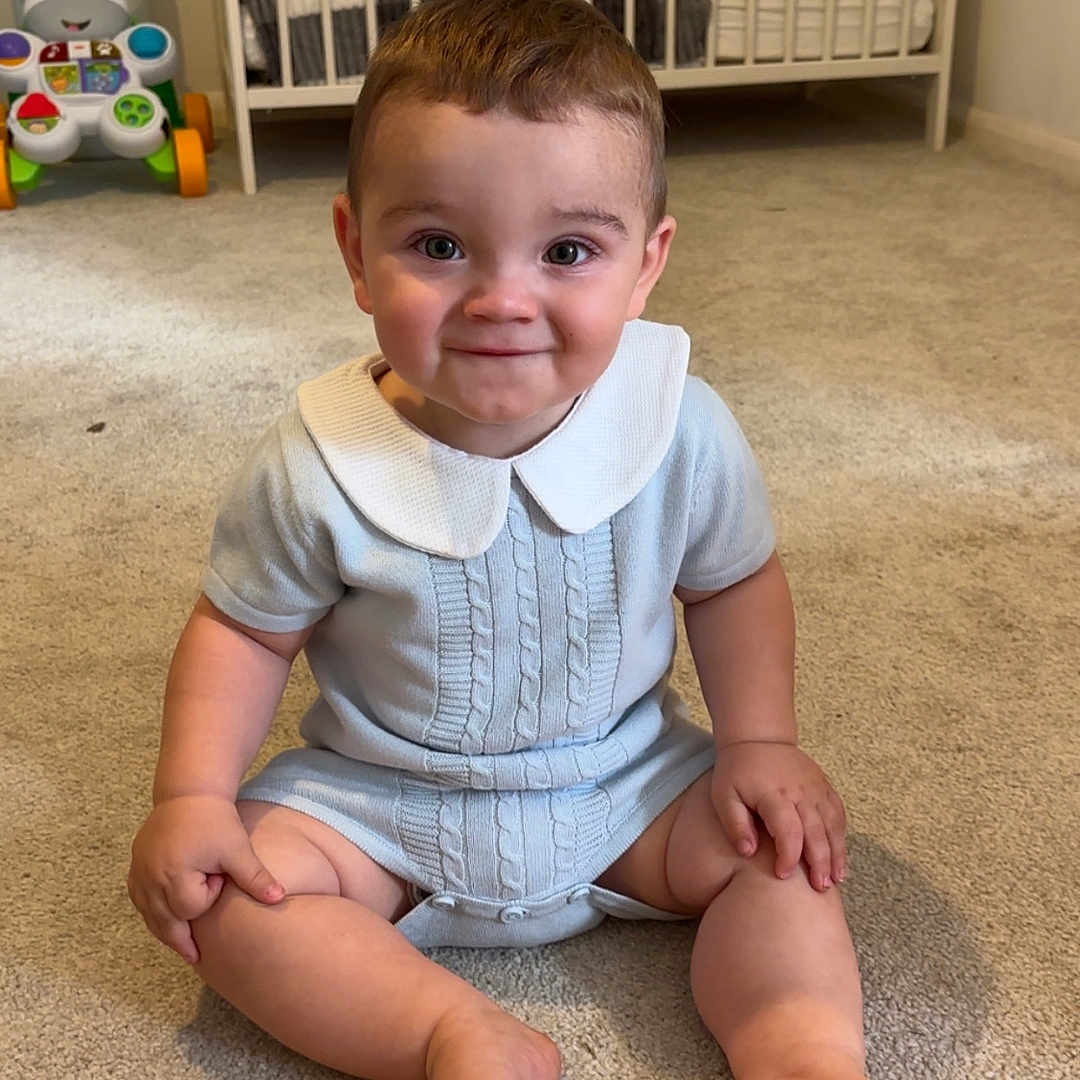 William is registered to the contest to win money with this photo: baby, carpet, child, clothing, crib, cute, face, floor, happy, head, home, indoor, nursery, person, playroom, romper, sitting, smiling, toddler, toy