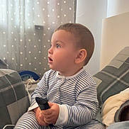 William joined the competition — help win amazing prizes! toddler, child, striped_pajamas, remote_control, indoor, curtains, polka_dots, blanket, pillow, bed, cozy, young_child, sitting, side_profile, curious, soft_lighting, home, casual, comfort, expression
