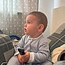 toddler, child, striped_pajamas, remote_control, indoor, curtains, polka_dots, blanket, pillow, bed, cozy, young_child, sitting, side_profile, curious, soft_lighting, home, casual, comfort, expression