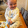 toddler, child, smiling, pajamas, table, fork, food, plate, fries, indoors, kitchen, wooden_table, happy, cute, clothing, person, face, sitting, home, playful