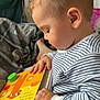 William is registered to the contest to win money with this photo: toddler, child, striped_pajamas, board_book, reading, indoor, closeup, side_profile, baby, hand, face, curly_hair, bed, blanket, foot, natural_light, cozy, cute, young_child, person