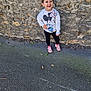 child, smiling, standing, outdoor, stone_wall, sweatshirt, mickey_mouse, minnie_mouse, pink_shoes, casual_clothing, young_girl, happy, sidewalk, playful, fashion, daylight, person, cute, joyful, urban