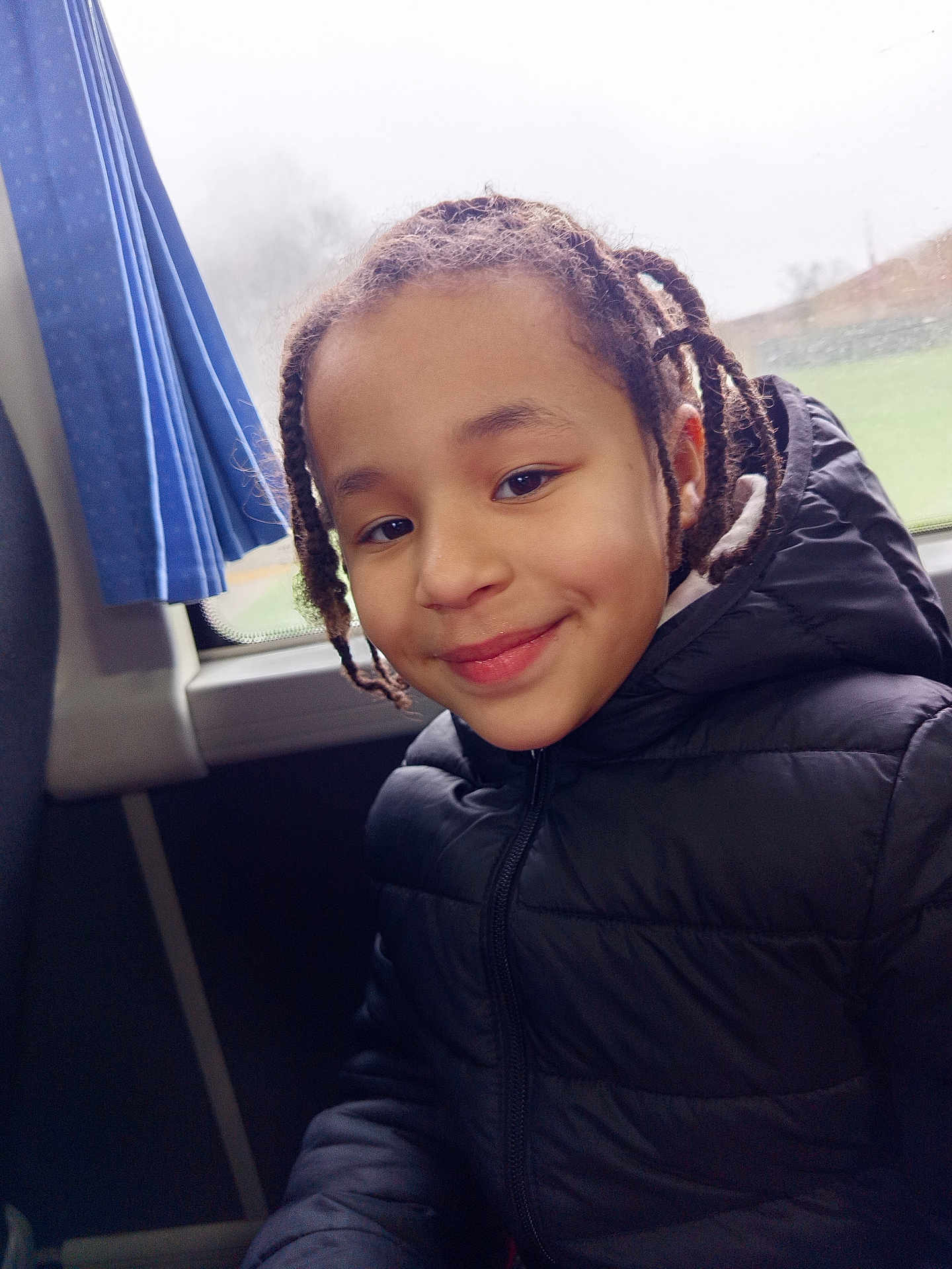 Felicia participe au concours pour gagner de l'argent avec cette photo : child, kid, smiling, braids, puffer_jacket, jacket, window, bus_seat, curtain, public_transport, portrait, happy, face, young_child, candid, close_up, seat, hair, hood, winter_clothing