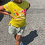 child, smiling, yellow_shirt, crochet_hat, beads, shorts, sneakers, parking_lot, sunny, shadow, sidewalk, bracelet, outdoor, person, casual, happy, standing, colorful, summer, fashion