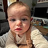 baby, child, face, indoor, living_room, television, plant, wooden_table, couch, blanket, clothing, portrait, curious, young_child, person, furniture, photo_frame, door, window, cozy