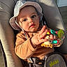 Elio participe au concours pour gagner de l'argent avec cette photo : baby, child, hat, striped_hat, brown_clothing, baby_seat, toy, giraffe_toy, hand, face, eyes, cushion, indoor, sunlight, curious, portrait, infant, seatbelt, closeup, cute