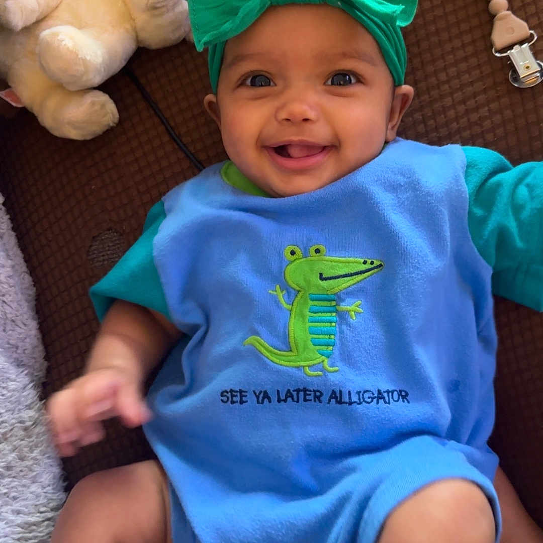 Willow is registered to the contest to win money with this photo: alligator_design, baby, baby_accessories, blue_clothing, brown_background, child, childhood, closeup, colorful, cute, happy, headband, indoors, infant, lying_down, playful, portrait, smiling, soft_texture, soft_toys