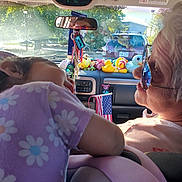 Arabella is registered to the contest to win money with this photo: child, elderly_person, car_interior, dashboard, toys, stuffed_animals, flag, purple_shirt, glasses, reflection, sunlight, window, seat, rearview_mirror, nature_outside, trees, road, license_plate, interaction, family