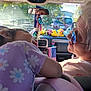 child, elderly_person, car_interior, dashboard, toys, stuffed_animals, flag, purple_shirt, glasses, reflection, sunlight, window, seat, rearview_mirror, nature_outside, trees, road, license_plate, interaction, family