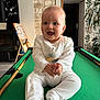 baby, child, smiling, white_clothing, onesie, pool_table, green_felt, indoor, stone_wall, plant, wooden_cue, furniture, room, joyful, clapping, happy, portrait, infant, sitting, playful