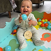 Côme participe au concours pour gagner de l'argent avec cette photo : baby, child, play_mat, toy_balls, blue_ball, smiling, indoor, floor, colorful, onesie, happy, infant, sitting, playtime, living_room, wooden_table, carpet, soft_toys, cute, young_child