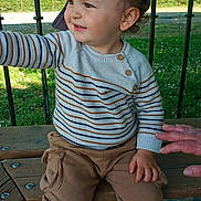 Elijah is registered to the contest to win money with this photo: toddler, child, outdoor, bench, cap, striped_sweater, curious, smiling, grass, fence, daylight, casual_clothing, person, brown_pants, black_shoes, hand, nature, playful, young_child, park