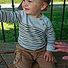 toddler, child, outdoor, bench, cap, striped_sweater, curious, smiling, grass, fence, daylight, casual_clothing, person, brown_pants, black_shoes, hand, nature, playful, young_child, park