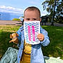 Elijah participe au concours pour gagner de l'argent avec cette photo : toddler, child, outdoor, park, blanket, plush_toy, card, grass, trees, bench, lamp_post, sky, lake, sunny, clothing, yellow_pants, blue_jacket, face, barefoot, person