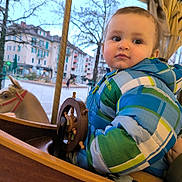 Elijah a rejoint le concours — aidez-le/la à gagner de superbes lots ! buildings, carousel, carousel_horse, child, city, daytime, face, fun, hand, outdoor, person, plaid_jacket, play, portrait, seat, toddler, toy, trees, winter_clothing, wooden_wheel