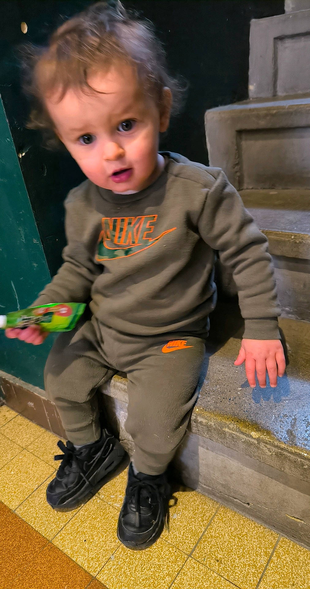 Elijah participe au concours pour gagner de l'argent avec cette photo : black_shoes, casual_clothing, child, concrete, curious, expression, face, floor_tiles, green_snack_pouch, hair, hand, indoor, looking, nike, portrait, sitting, stairs, toddler, tracksuit, young_child